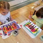 Dzieci siedzą przy stoliku, malują farbami tło do pracy plastycznej. Praca przedstawia jesienne drzewa.