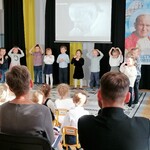 Dzieci z grupy 2 występują przez zebraną publicznością. W tle na ekranie i  na banerze widać sylwetkę Papieża Jana Pawła II..jpg