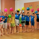 Zdjęcie przedstawia grupę dzieci ustawionych w rzędzie z piłkami uniesionymi nad głową.jpg
