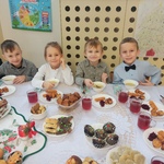 Dzieci z grupy Pszczółki siedzą przy Wigilijnym stole. Stół jest przyozdobiony białym obrusem, świecznikami i stroikiem świątecznym. Na stole są również  przyniesione przez dzieci potrawy Wigilijne. jpg