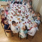 Dzieci z grupy Pszczółki siedzą przy Wigilijnym stole. Stół jest przyozdobiony białym obrusem, świecznikami i stroikiem świątecznym. Na stole są również  przyniesione przez dzieci potrawy Wigilijne .jpg