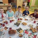 Dzieci z grupy Pszczółki siedzą przy Wigilijnym stole. Stół jest przyozdobiony białym obrusem, świecznikami i stroikiem świątecznym. Na stole są również  przyniesione przez dzieci potrawy Wigilijne .jpg