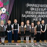 Na zdjęciu widać dzieci z najstarszej. Stoją w szeregu przed kurtyną z napisem i dekoracją kwiatową. Śpiewają piosenkę .jpg
