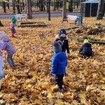 Na zdjęciu widać dzieci z najmłodszej grupy. Dzieci bawią się wśród jesiennych liści. Na dalszym planie zobaczyć można wybrane sprzęty terenowe znajdujące się na placu przedszkolnym.jpg