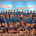 Dzieci z całego przedszkola pozują do pamiątkowego zdjęcia. W tle widać napis  - DZIEŃ PRZEDSZKOLAKA