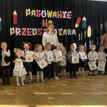Pasowanie na Przedszkolaka .Dzieci z I grupy  podczas pamiątkowego zdjęcia z p. dyrektor i p. Ulą. W tle na czarnej kurtynie widać napis PASOWANIE NA PRZEDSZKOLAKA i dekorację - kolorowe kredki.