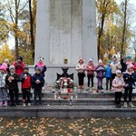 Dzieci z grupy Pszczółek stoją ze zniczami w rękach przed centralnym pomnikiem z napisem CZEŚĆ POLEGŁYM  i wmurowanym metalowym Krzyżem Virtuti Militari.
