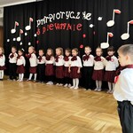Dzieci z II gr. w strojach galowych podczas recytacji wiersza - Katechizm polskiego dziecka. W tle czarna kurtyna dekoracją i napisem  - Patriotyczne śpiewanie.