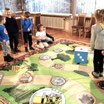 Dzieci grają w żywą grę planszową utrwalającą wiadomości o Polsce. Dwie dziewczynki są pionkami poruszającymi się po planszy, pozostałe dzieci rzucają dużą kostką i odpowiadają na pytania zadawane przez Panią Anię.