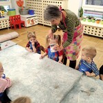 Grupa dzieci siedzi przy stolikach. Osoba prowadząca nakłada z pojemnika szlam.